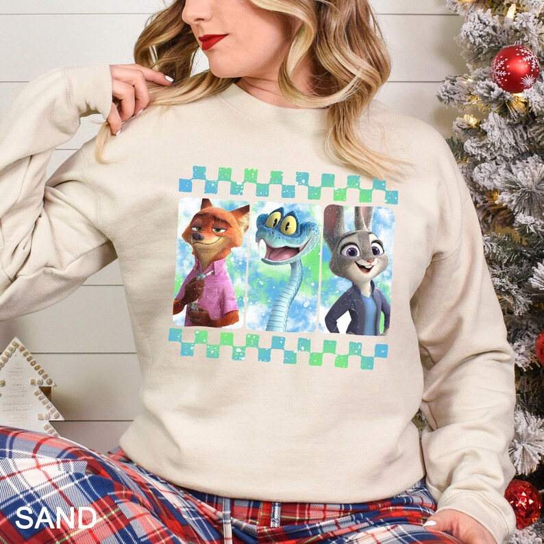 May include: Beige sweatshirt featuring three cartoon characters: a fox in a pink shirt, a blue snake, and a rabbit in a blue jacket. The design is framed with a blue and green checkered pattern.