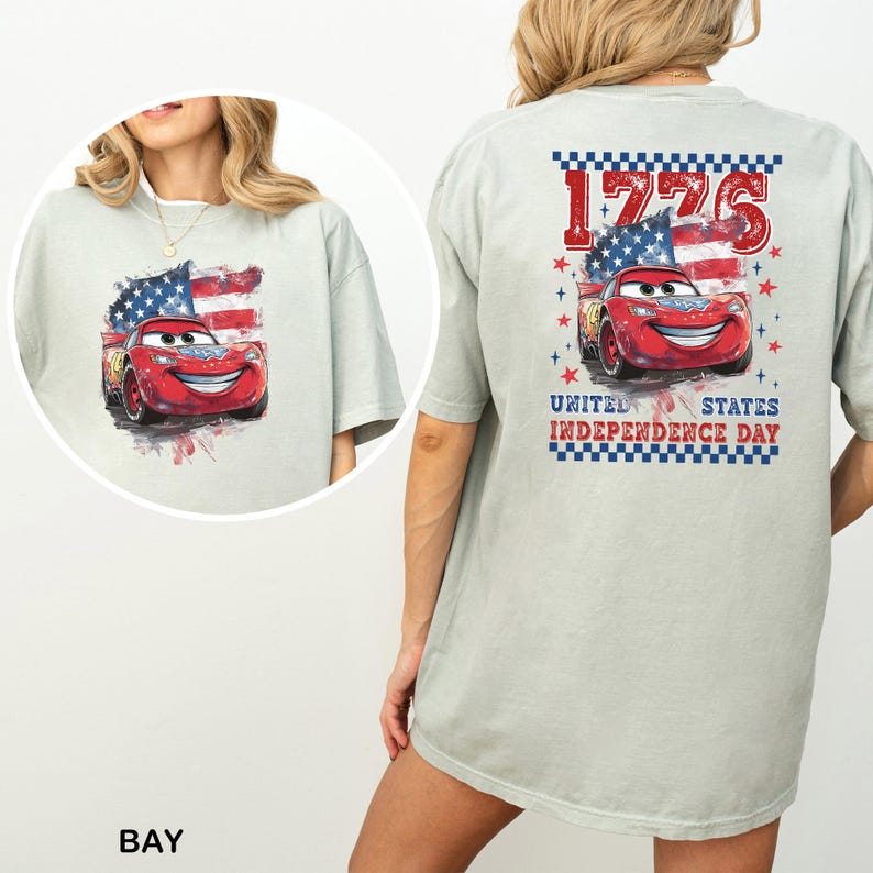 May include: A light grey t-shirt featuring a graphic of a red race car with a smiling face, set against a stylised American flag. The back design includes the text 1776 and UNITED STATES INDEPENDENCE DAY, framed by a blue and white chequered border.