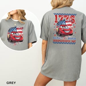 May include: A grey t-shirt featuring a graphic of a red race car with a smiling face, positioned in front of an American flag. The back design includes the text 1776 and United States Independence Day, framed by a blue and white checkered border.