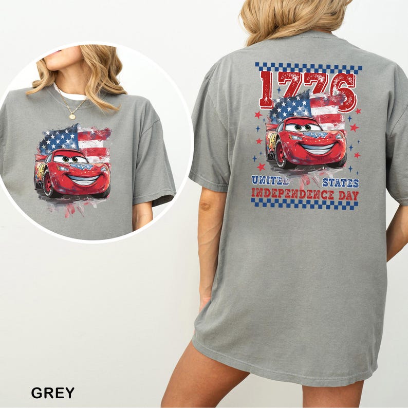 May include: A grey t-shirt featuring a graphic of a red race car with a smiling face, positioned in front of an American flag. The back design includes the text 1776 and United States Independence Day, framed by a blue and white checkered border.