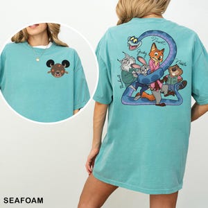 May include: Seafoam green t-shirt featuring a cartoon graphic on the front and back. The front has a brown bear with black ears. The back has a blue snake with cartoon characters and the names Judy, Nick, and Nibbles.