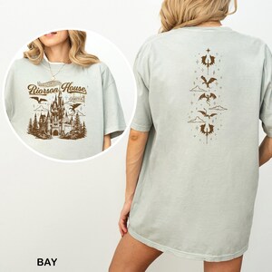 May include: A light green t-shirt with a brown graphic of a castle, trees, and dragons. The text "Riorrson House" is above the castle. The back of the shirt has a vertical design of dragons and clouds.
