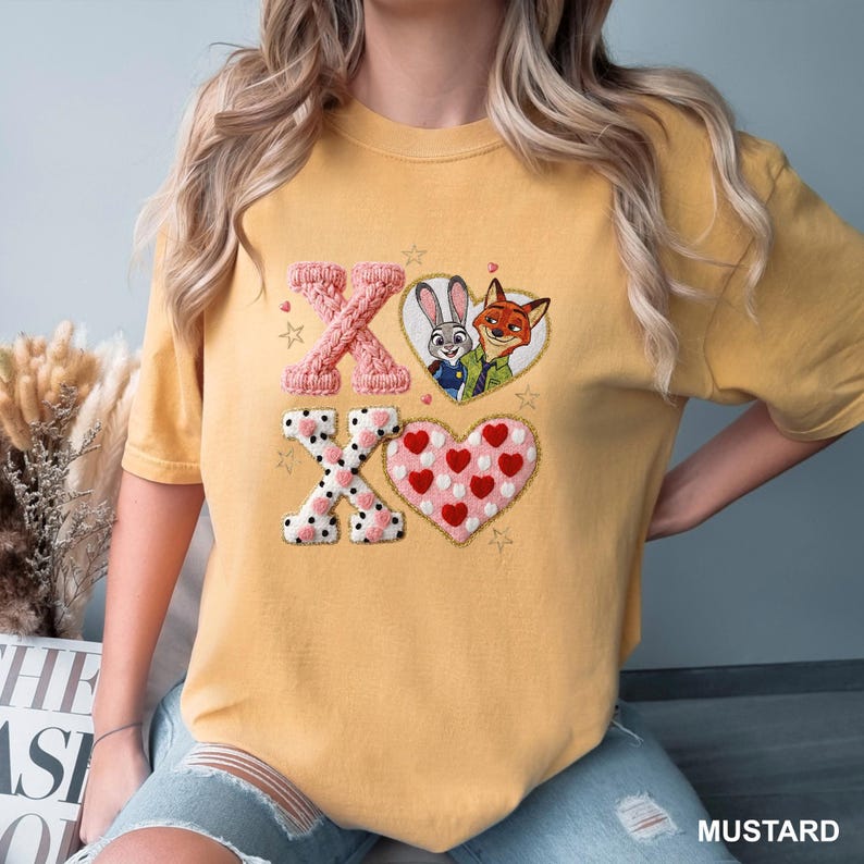 Może przedstawiać: Musztardowa koszulka z literami "XO" w dekoracyjnym wzorze. "X" jest r&oacute;żowe, a "O" ma kształt serca z kresk&oacute;wkowym wizerunkiem Judy Hopps i Nicka Wilde'a. Dolne "X" jest białe z czarnymi kropkami, a serce jest r&oacute;żowe z czerwonymi sercami.
