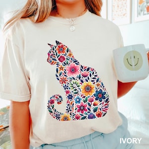 May include: Ivory-colored t-shirt featuring a floral cat design. The cat silhouette is filled with colorful flowers in shades of pink, yellow, blue, and orange. The word "IVORY" is printed at the bottom of the shirt.