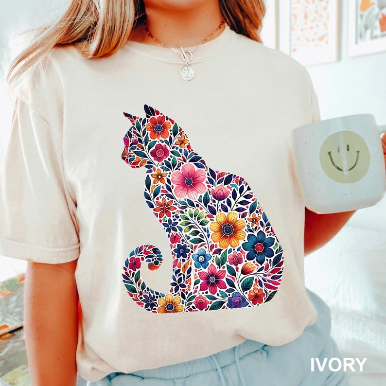 May include: Ivory-colored t-shirt featuring a floral cat design. The cat silhouette is filled with colorful flowers in shades of pink, yellow, blue, and orange. The word "IVORY" is printed at the bottom of the shirt.
