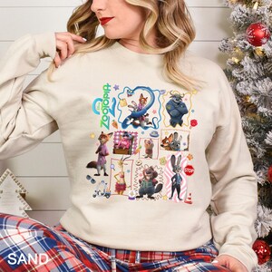 May include: A sand-coloured sweatshirt with a colourful graphic of characters from the Disney film "Zootopia." The graphic includes images of a fox, a bunny, and other animal characters. The word "Zootopia" is written in green and blue script.
