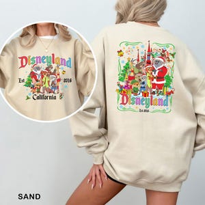 May include: Sand-colored sweatshirt with a Disneyland graphic on the front and back. The front graphic says "Disneyland California Est. 2016" with cartoon characters. The back graphic has a similar design with a castle and Santa.
