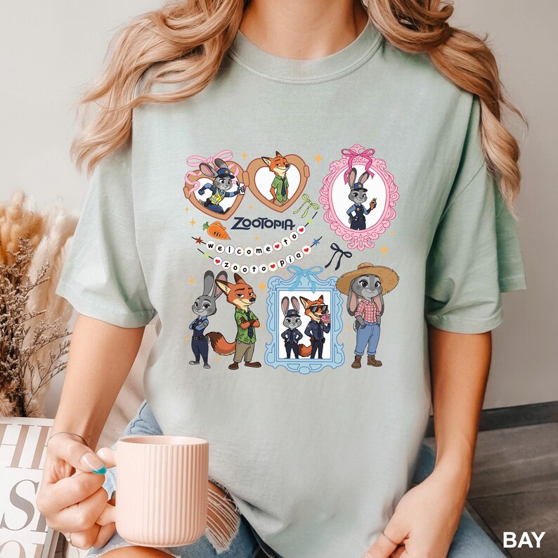 Op de afbeelding: Lichtgroen T-shirt met een Zootopia-ontwerp. Het ontwerp bevat stripfiguren zoals een vos en een konijn, in kaders en harten. De woorden "Zootopia" en "welcome to zootopia" staan op het T-shirt.