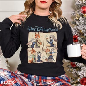 Puede incluir: Sudadera negra con un dise&ntilde;o de dibujos animados de cuatro paneles con personajes de Zootopia. El panel superior dice "Walt Disney World". La esquina inferior izquierda de la sudadera tiene la palabra "BLACK".