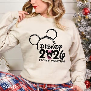 以下が含まれることがあります： 黒いミッキーマウスの頭のデザインと、黒とピンクの「DISNEY 2026 FAMILY VACATION」の文字が入った、サンドカラーのスウェットシャツ。スウェットシャツは長袖です。