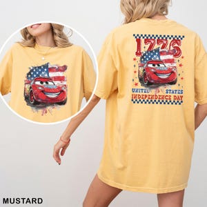 May include: A mustard yellow t-shirt featuring a graphic of a red race car with eyes on the windshield, set against a stylised American flag. The back print includes the text 1776 and UNITED STATES INDEPENDENCE DAY with red stars.