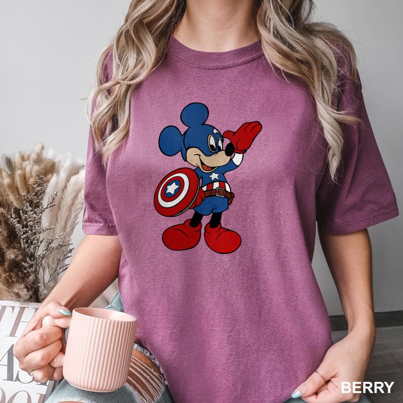 May include: Berry-colored t-shirt featuring a cartoon Mickey Mouse dressed as Captain America, holding a shield. The character is in blue, red, and white. The shirt has short sleeves and a crew neck. The word "BERRY" is printed at the bottom right.