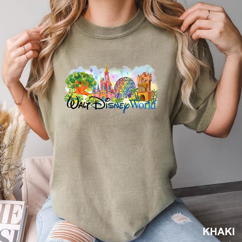 May include: A khaki-colored crew neck t-shirt featuring a colorful graphic print of the Walt Disney World skyline, including the Tree of Life, Cinderella Castle, Spaceship Earth, and the Tower of Terror, with the text Walt Disney World below.