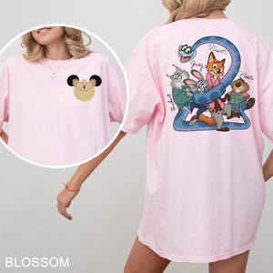 May include: Light pink t-shirt with a cartoon graphic on the front and back. The front features a mouse-like character, while the back displays a blue snake and cartoon characters. The word "Blossom" is printed at the bottom.