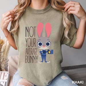 May include: Khaki t-shirt with a cartoon bunny in a police uniform, and the text "NOT YOUR AVERAGE BUNNY". The bunny has pink ears and is holding a briefcase. The shirt is a relaxed fit. The colour is khaki.