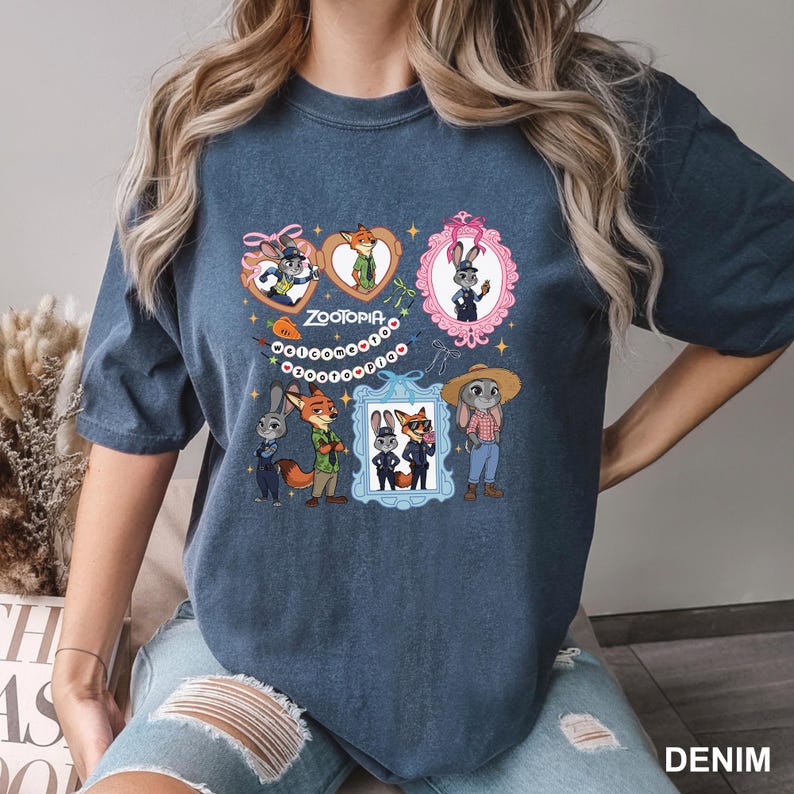 Op de afbeelding: Denim blauw t-shirt met een kleurrijk grafisch ontwerp met personages uit de animatiefilm "Zootopia". Het ontwerp bevat ingelijste portretten en hartvormige elementen met het woord "Zootopia" in het midden. Het shirt is gemaakt van zacht materiaal.