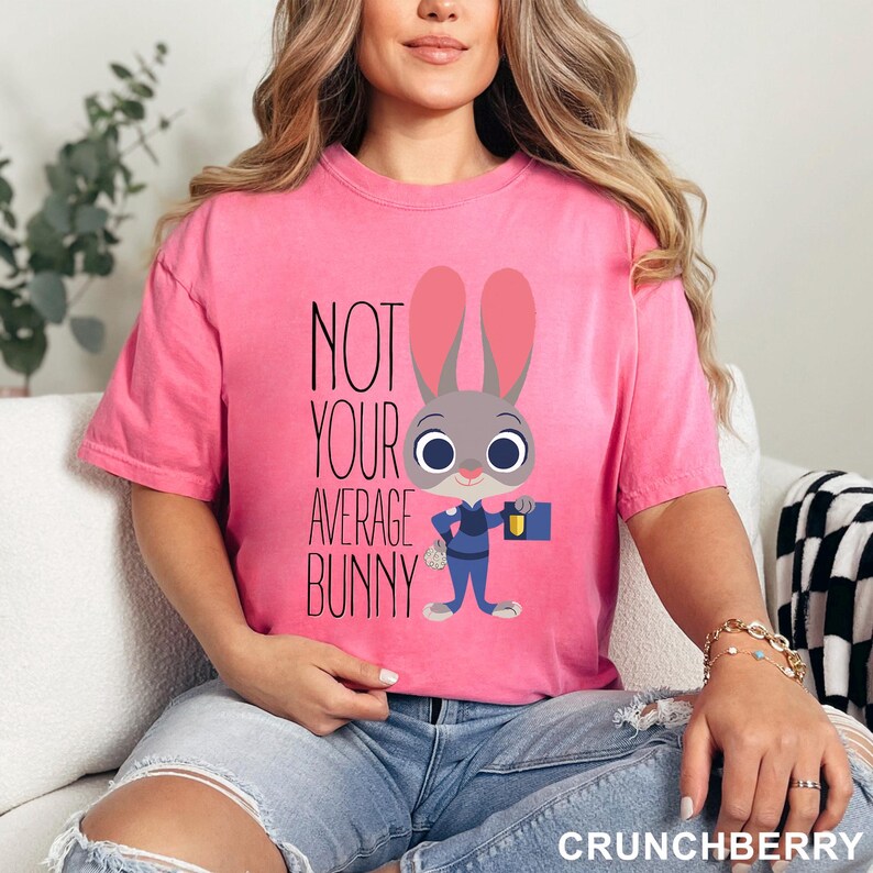 May include: A pink t-shirt featuring the phrase "NOT YOUR AVERAGE BUNNY" and a cartoon bunny in a police uniform, carrying a briefcase. The t-shirt is a solid pink colour.