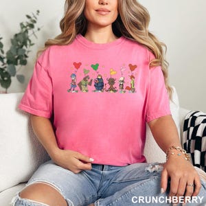 May include: A hot pink t-shirt featuring a cartoon graphic of several whimsical characters holding heart-shaped balloons. The word "CRUNCHBERRY" is printed in white at the bottom of the image. The shirt is made of a soft material.