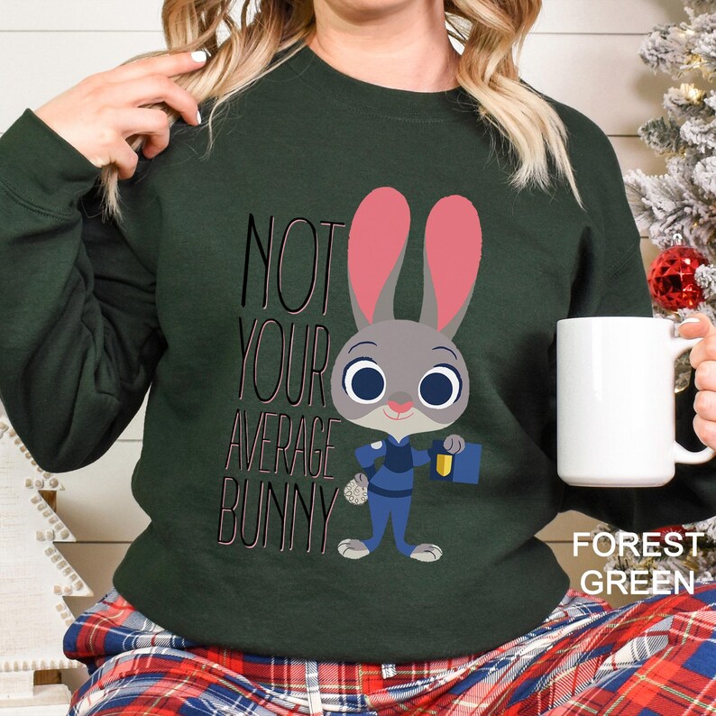Op de afbeelding: Bostgroen sweatshirt met de tekst "NOT YOUR AVERAGE BUNNY" en een cartoon konijn karakter in een politie-uniform. Het konijn heeft roze oren en houdt een badge en een blauwe tas vast. Een witte mok wordt in de hand gehouden.