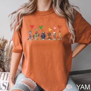 May include: Burnt orange t-shirt featuring a whimsical cartoon design of several characters, each holding a heart-shaped balloon. The characters are diverse in appearance, with varying colors and outfits. The shirt has short sleeves and a relaxed fit. The word "YAM" is printed in white at the bottom right.
