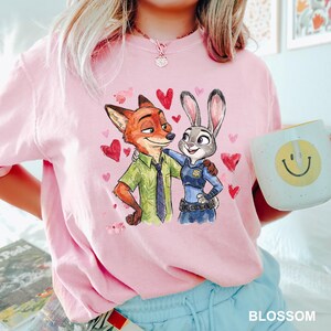 Może przedstawiać: R&oacute;żowa koszulka z akwarelową ilustracją Nicka Wilde'a i Judy Hopps z Zootopii, otoczona czerwonymi sercami. Koszulka ma okrągły dekolt i kr&oacute;tkie rękawy. Na zdjęciu trzymany jest biały kubek z ż&oacute;łtą uśmiechniętą buzią.