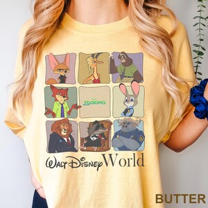 May include: Butter-colored t-shirt featuring a grid of Zootopia characters. The shirt includes the text "Zootopia" and "Walt Disney World" in black. The shirt is made of a soft material.