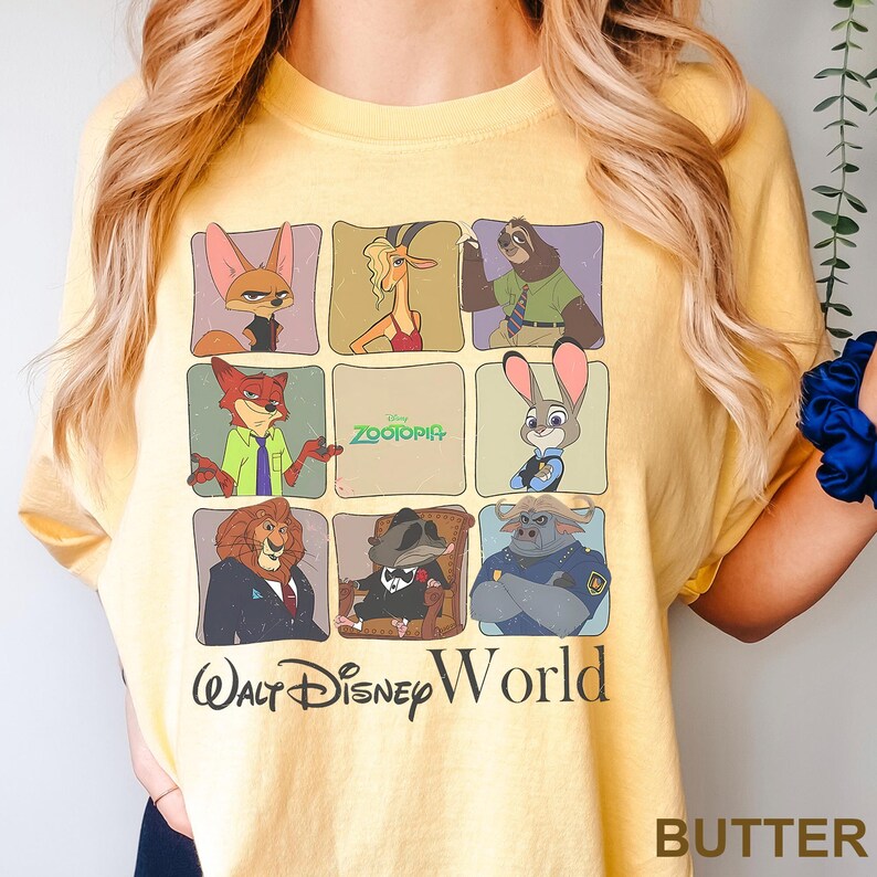 May include: Butter-colored t-shirt featuring a grid of Zootopia characters. The shirt includes the text "Zootopia" and "Walt Disney World" in black. The shirt is made of a soft material.