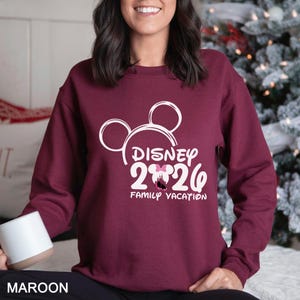 以下が含まれることがあります： ミッキーマウスの耳、テキスト「DISNEY 2026」、および「FAMILY VACATION」を備えた白いディズニーのグラフィックが特徴の、マルーンのスウェットシャツ。スウェットシャツは無地のマルーン色です。