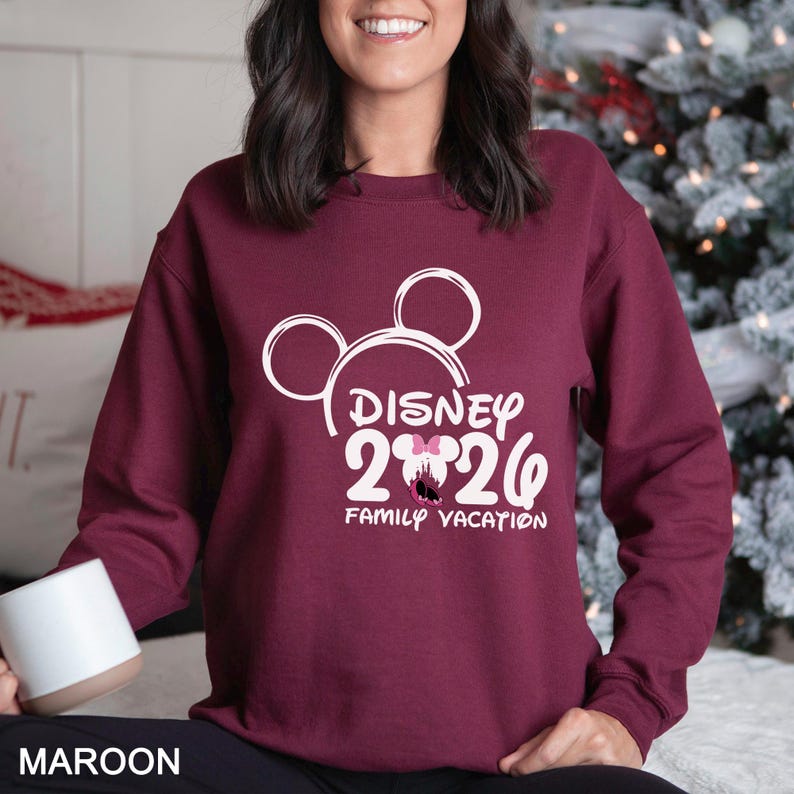以下が含まれることがあります： ミッキーマウスの耳、テキスト「DISNEY 2026」、および「FAMILY VACATION」を備えた白いディズニーのグラフィックが特徴の、マルーンのスウェットシャツ。スウェットシャツは無地のマルーン色です。