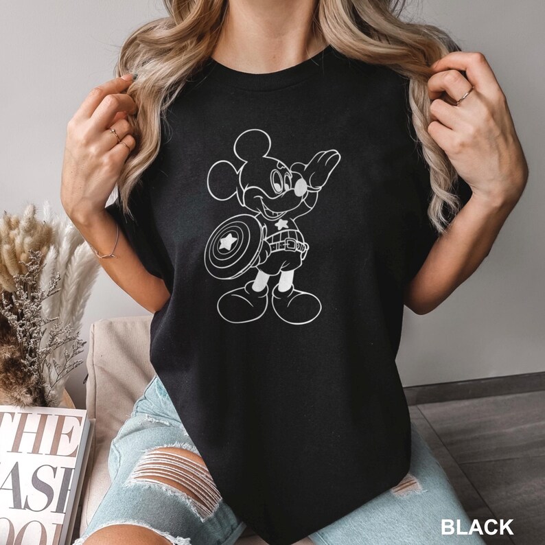 May include: Black t-shirt featuring a white graphic of Mickey Mouse dressed as Captain America, holding a shield. The text "BLACK" is at the bottom right. The shirt is a crew neck.
