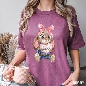 May include: Berry-colored t-shirt featuring a cartoon bunny holding a heart-shaped sign with the word "Love." The bunny wears a pink bow and a blue uniform. The shirt is a solid color with a crew neck.