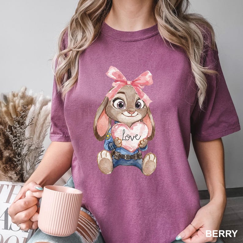 May include: Berry-colored t-shirt featuring a cartoon bunny holding a heart-shaped sign with the word "Love." The bunny wears a pink bow and a blue uniform. The shirt is a solid color with a crew neck.