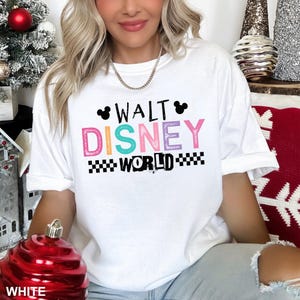May include: A white short-sleeved t-shirt featuring the text WALT DISNEY WORLD in colorful, textured lettering. The design includes two black Mickey Mouse ear silhouettes above the word WALT and black checkered patterns flanking the word WORLD.
