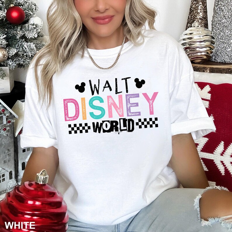 May include: A white short-sleeved t-shirt featuring the text WALT DISNEY WORLD in colorful, textured lettering. The design includes two black Mickey Mouse ear silhouettes above the word WALT and black checkered patterns flanking the word WORLD.
