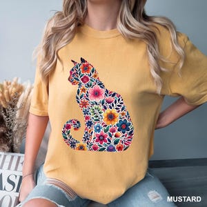 May include: Mustard-colored t-shirt featuring a floral cat design. The cat silhouette is filled with colorful flowers, leaves, and intricate details. The word "MUSTARD" is printed in white at the bottom right corner of the image.