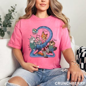 May include: A pink t-shirt featuring a cartoon graphic of several animal characters, including a fox, rabbit, and beaver, with the number 2 and the word "Crunchberry" at the bottom. The characters are set against a blue snake-like design.