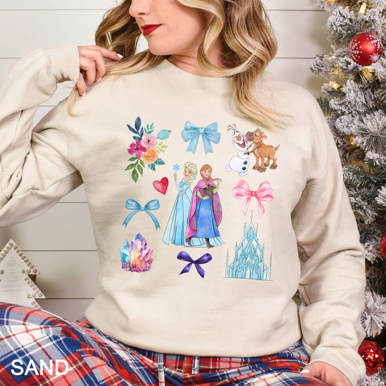以下が含まれることがあります： 人気の漫画映画のキャラクター、弓、花、城のイラストが描かれたサンドカラーのスウェットシャツ。デザインにはハート、クリスタル、雪だるまが含まれています。「SAND」の文字が下部に印刷されています。
