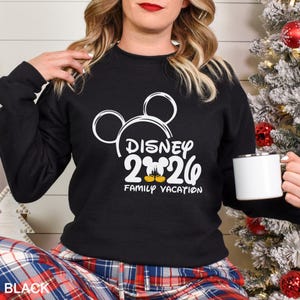 以下が含まれることがあります： 白のディズニー2020グラフィックと「Family Vacation」の文字が入った黒のクルーネックのトレーナー。トレーナーにはミッキーマウスの耳が付いています。「BLACK」の文字が左下に印刷されています。