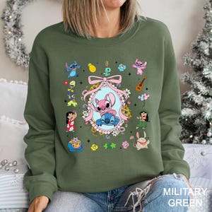 May include: Military green sweatshirt featuring a colorful graphic of Stitch and Angel characters. The design includes a central oval frame with the characters, surrounded by floral and character illustrations. The sweatshirt is a long-sleeved crew neck.