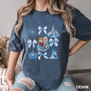 May include: Denim blue t-shirt featuring a graphic of characters and elements from the animated film. The design includes images of Anna, Elsa, Olaf, bows, a castle, a snowflake, and an ice structure. The word "DENIM" is printed on the bottom right.