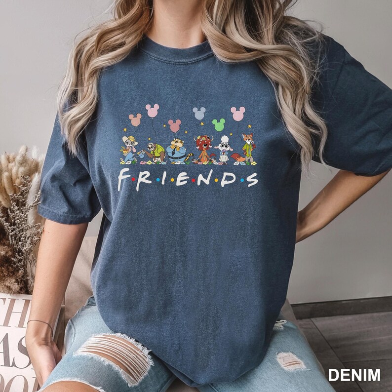 May include: A denim blue t-shirt featuring a colorful graphic of cartoon characters and the word "FRIENDS" in a playful font. Above the characters are Mickey Mouse head silhouettes in various colors. The shirt is a casual, comfortable style.