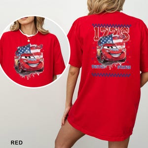 May include: A bright red t-shirt featuring a graphic of a red race car with eyes on the windscreen, set against a stylised American flag. The back design includes the text 1776 and UNITED STATES, framed by a blue and red chequered border.