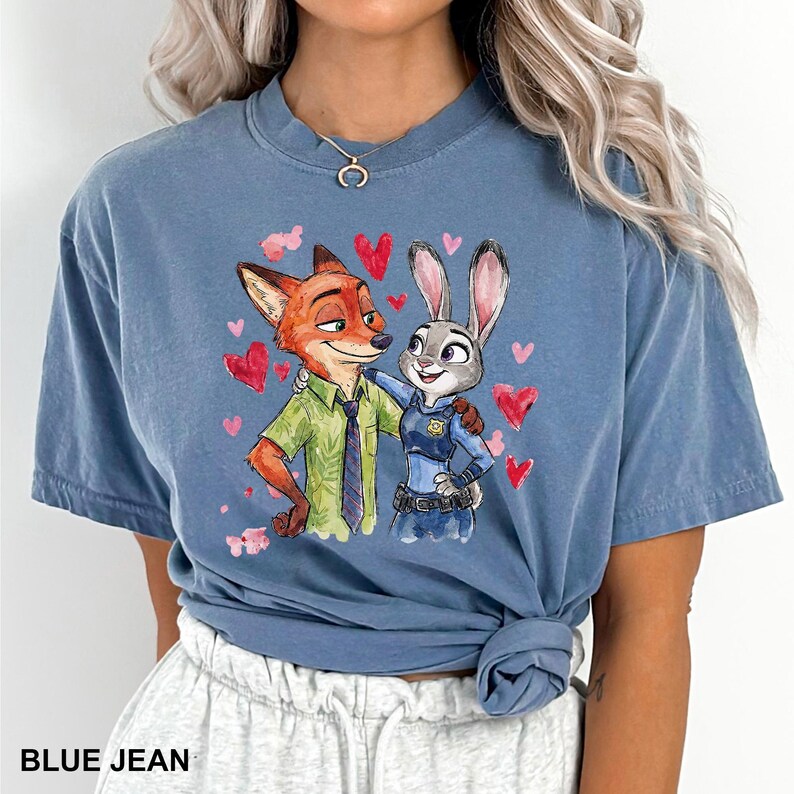 Może przedstawiać: Niebieska koszulka dżinsowa z kresk&oacute;wkową ilustracją Nicka Wilde'a i Judy Hopps z Zootopii, otoczona czerwonymi sercami. Koszulka ma okrągły dekolt i kr&oacute;tkie rękawy. Tekst "BLUE JEAN" jest wydrukowany na dole.