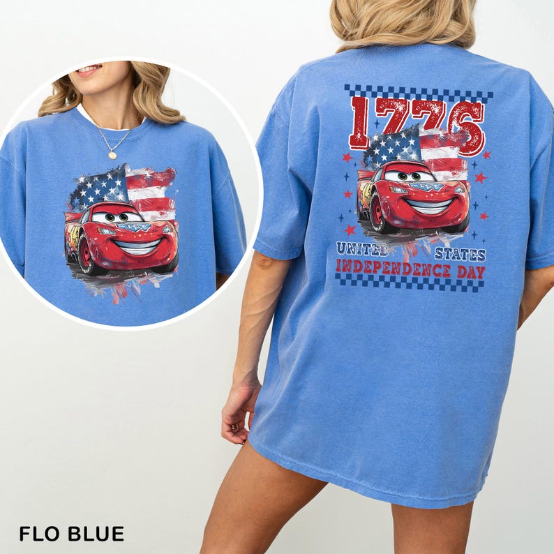 May include: A light blue t-shirt featuring a graphic of a red race car with a smiling face in front of an American flag. The back design includes the text 1776 and United States Independence Day with red stars and a blue checkered border.