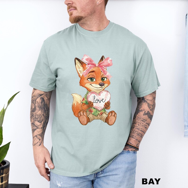 May include: A light green t-shirt featuring a cartoon fox holding a heart-shaped sign that says "love". The fox has orange fur, a pink bow, and is wearing a Hawaiian shirt. The word "BAY" is printed in the bottom right corner.