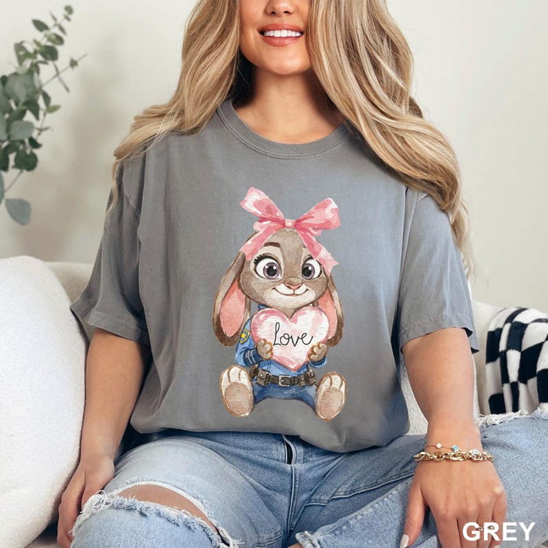 May include: A grey t-shirt featuring a cartoon bunny holding a heart-shaped sign with the word "Love". The bunny has a pink bow and is wearing a blue uniform. The shirt is a solid grey color.