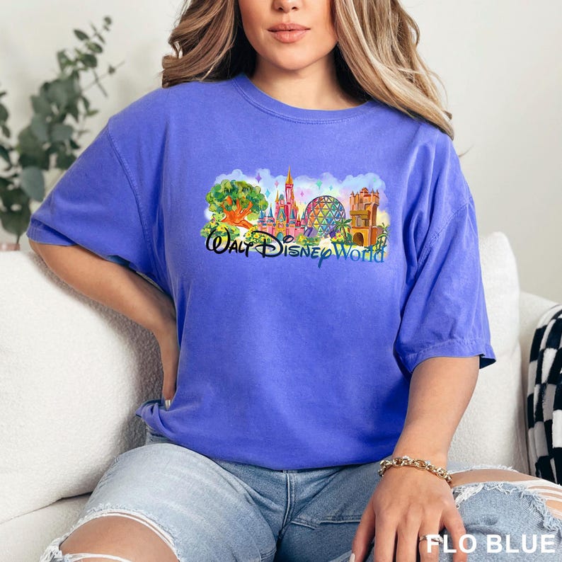 May include: A periwinkle blue short-sleeve t-shirt featuring a colorful graphic print of the Walt Disney World theme park landmarks, including the Tree of Life, Cinderella Castle, Spaceship Earth, and the Tower of Terror with the text Walt Disney World.
