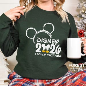 以下が含まれることがあります： 白いミッキーマウスの耳と「Disney 2026 Family Vacation」の文字が入ったフォレストグリーンのスウェットシャツ。白いマグカップが手に持たれています。スウェットシャツは単色です。