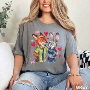 Może przedstawiać: Szara koszulka z akwarelową ilustracją Nicka Wilde'a i Judy Hopps z Zootopii, otoczona r&oacute;żowymi sercami. Nick ma na sobie zieloną koszulę i krawat, a Judy jest w swoim policyjnym mundurze. Słowo "GREY" jest wydrukowane na dole.