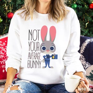 Op de afbeelding: Witte sweatshirt met een cartoon konijn in politie-uniform en de tekst "NOT YOUR AVERAGE BUNNY". Het konijn is grijs met roze oren en blauwe ogen. De sweatshirt is een crew neck stijl.